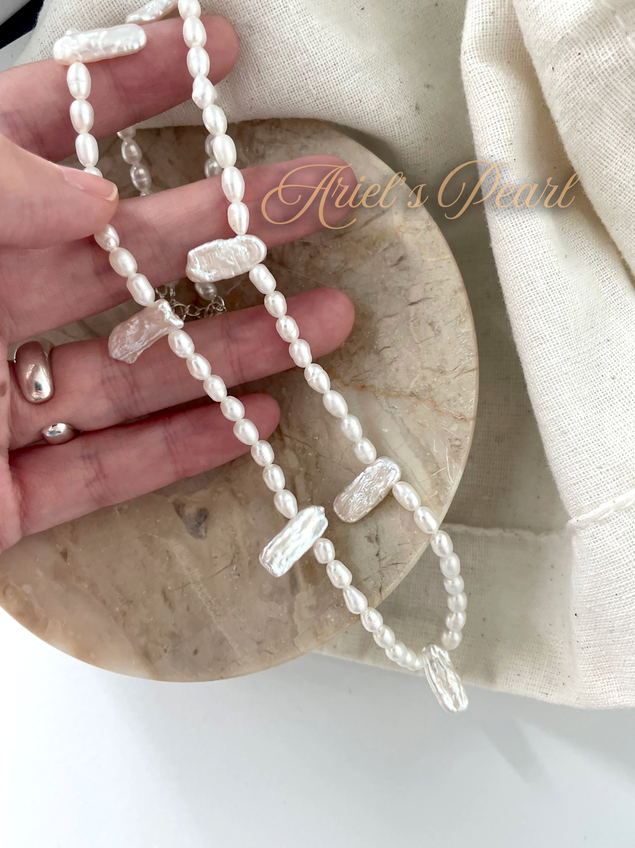 Pearl necklace held by a hand on a wooden surface with 'Ariel's Pearl' branding.