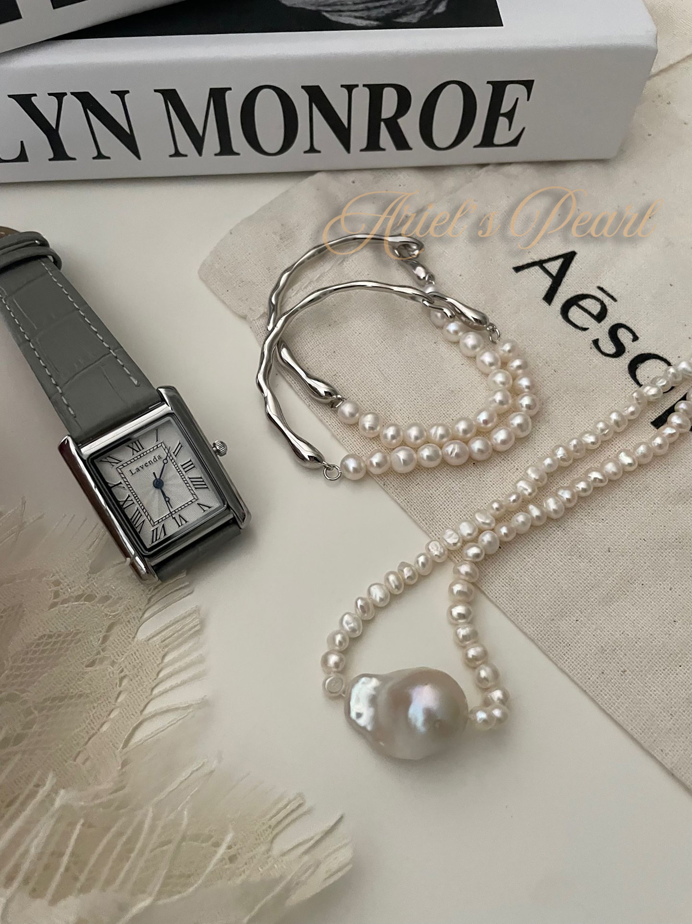 Watch and pearl necklace on a textured surface with 'Aesop' branding.