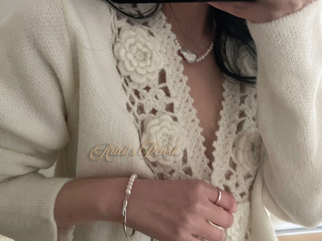 Close-up of a person wearing a cream-colored lace top with a brand name, pearl bracelet, and ring.