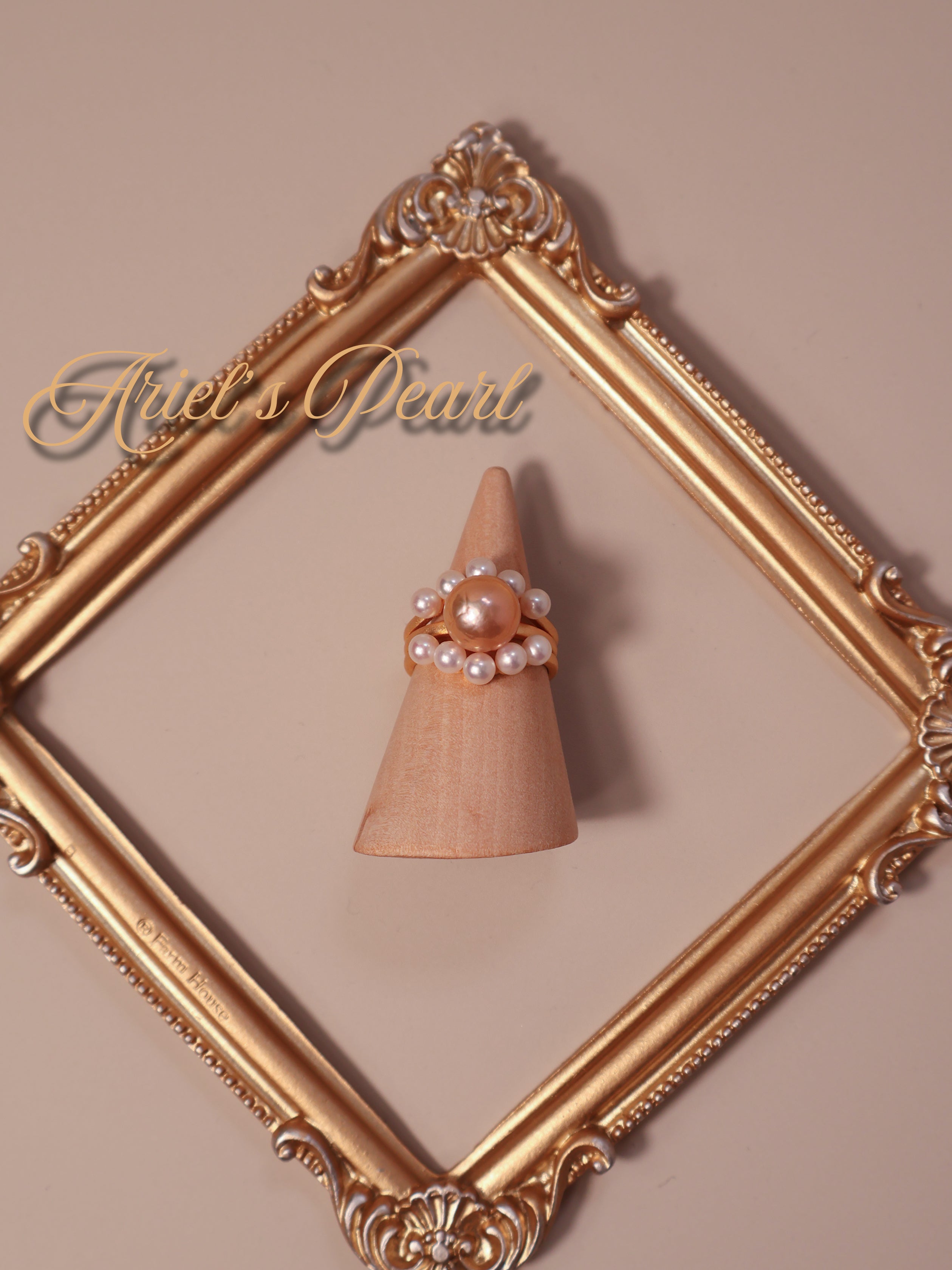 Ring with pearls on a wooden stand inside an ornate gold frame, branded 'Ariel's Pearl'.