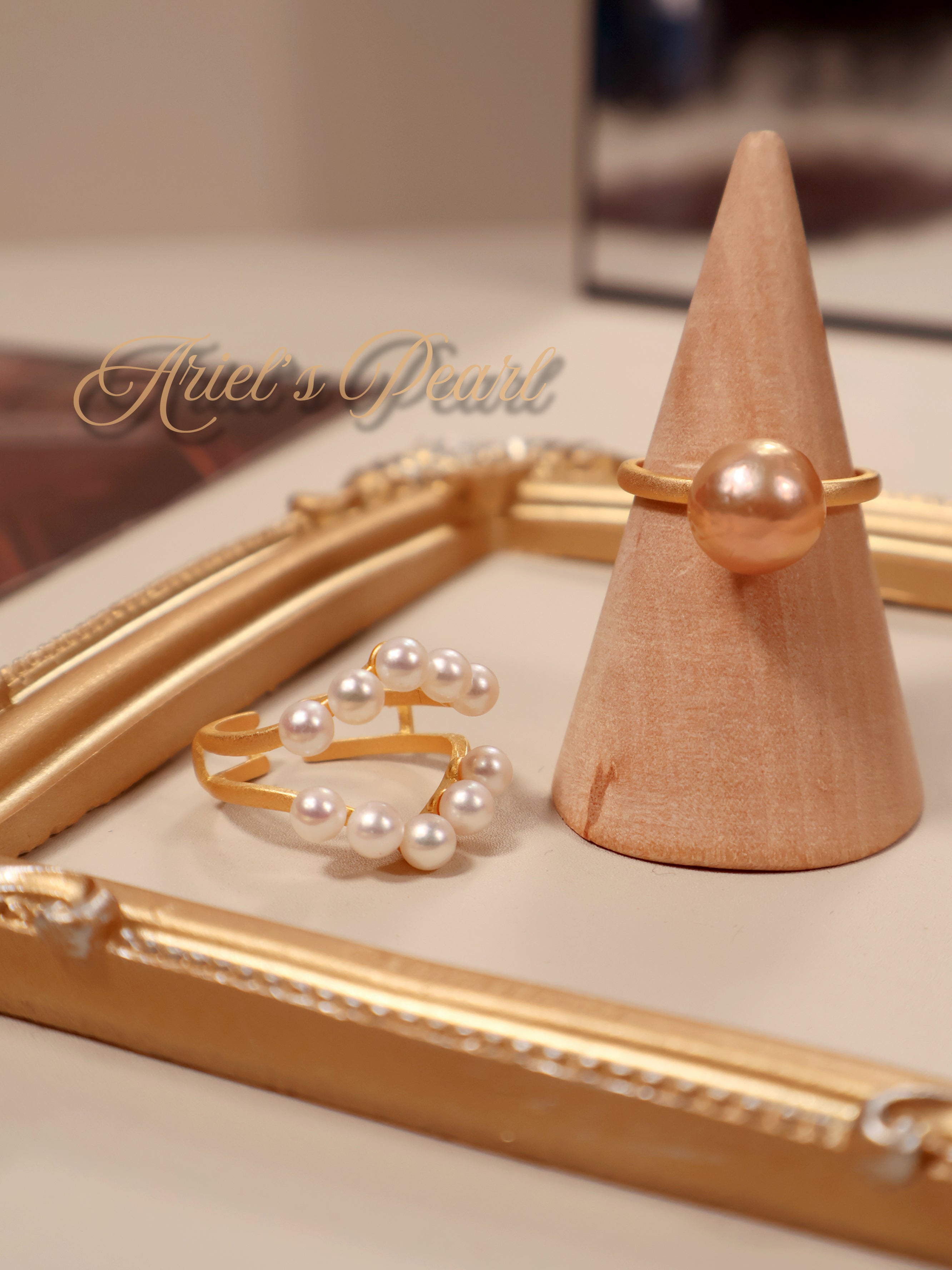 Gold and pearl rings on a wooden stand with 'Ariel's Pearl' branding.